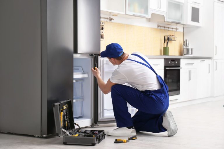 A man in overalls repairs a refrigerator, demonstrating appliance repair skills in a home setting.
